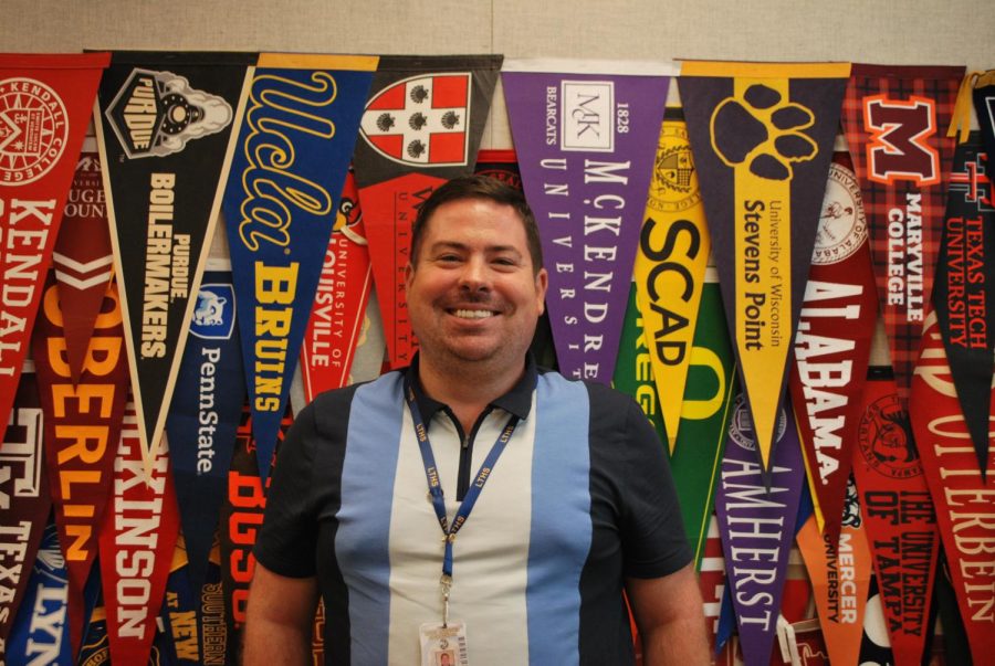 Cody Dailey poses against the college flag wall in the College/Career Center, where he has taken over the position of the College Coordinator (Mardegan/LION).