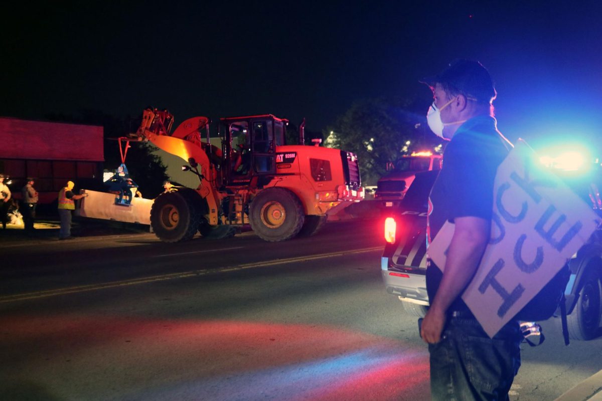 Protester watches barricades be placed bordering sidewalks to keep streets clear of cars and people with a poster in hand (Brooks/LION).
